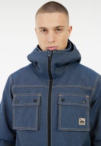 Junger Mann mit kurzen Haaren, der eine blaue Jeansjacke mit zwei vorderen Knopf-Taschen und einem hohen Reißverschlusskragen trägt.