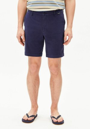 Uomo che indossa pantaloncini blu navy, maglietta a righe e infradito blu navy, in piedi con le braccia rilassate lungo i fianchi su sfondo bianco.