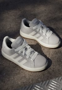 Světlé šedé tenisky vyrobené z texturované suede s bílými pruhy, mají kulatou špičku a gumovou podrážku. Šňůrky odpovídají barvě bot.