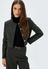 Grijze korte jas met een gestructureerde afwerking, voorzien van gouden knoopaccenten en een voorzak, gecombineerd met een zwart high-neck top en op maat gemaakte broeken.