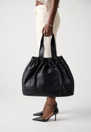 Person wearing cream pants and black high heels holds large black leather tote bag with external pocket.