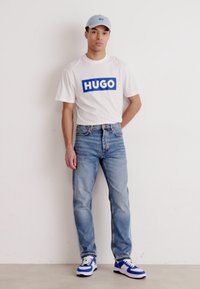 Tricou alb cu logo-ul albastru "HUGO", șapcă albastră deschisă, blugi albaștri deschiși și adidași albi cu accente albastre. Stând lângă un perete neutru.