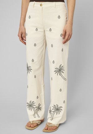 Pantalon écru avec motifs noirs de palmiers et de gouttes, bouton à l'avant, porté avec des sandales beiges et un haut marron.