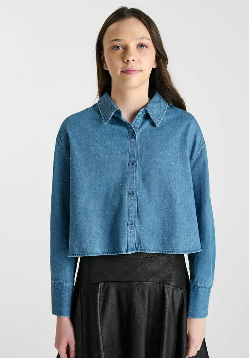 Korte spijkerblouse in lichtblauw met subtiele stoftextuur en twee voorzakken, gecombineerd met een zwarte leren rok.