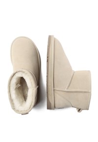 Beige suede ankle boots with a soft, fluffy interior, flat rubber soles, and stitched detailing. Features a pull tab on the back.
