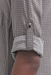 Manche d'une chemise à motifs avec de petits losanges noirs et marron clair, retroussée et maintenue par une patte boutonnée sur un bras à la peau foncée.