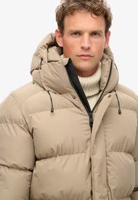 Giacca puffer beige con cappuccio, dotata di chiusura con zip, accenti neri e texture trapuntata, indossata sopra un maglione a collo alto crema.