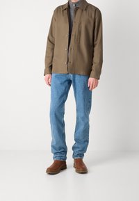 Chaqueta marrón de botones sobre una camisa gris, combinada con pantalones de mezclilla azules y botas marrones. Incluye un bolsillo en el pecho y una textura acanalada.