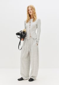 Graues Langarmoberteil mit Knopfdetails, kombiniert mit weiten grauen Hosen. Das Model hält eine kleine schwarze Handtasche und trägt schwarze Schuhe.