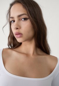 Primo piano di una persona con lunghi capelli castani, che indossa una maglietta bianca aderente con un ampio scollo, pelle liscia e colore delle labbra neutro.