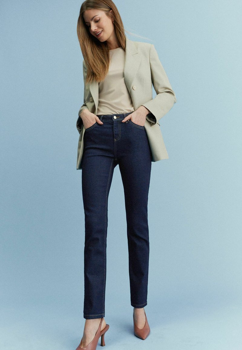 Mörkblå slim-fit jeans med kontrasterande sömmar, i kombination med en ljusbeige topp och en ljusgrön kavaj. Bruna klackskor fulländar looken.