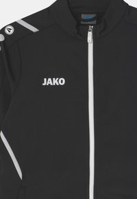 Zwarte sportjack gemaakt van gladde stof met een witte rits, een 'JAKO' logo en reflecterende accenten op de schouders.