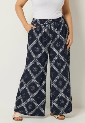 Kvinde iført marineblå bukser med vide ben med hvide geometriske blomsterprint, elastisk talje og sidelommer, kombineret med hvid top og sandaler.