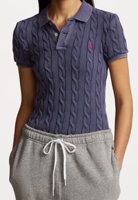 Lila kabelstickad pikétröja med krage och treknappslinning, kombinerad med grå tights med dragsko och sidfickor.