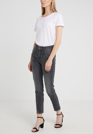 Frau, die ein weißes T-Shirt mit kurzen Ärmeln, hoch taillierte dunkelgraue Jeans mit vorderen Nähten und schwarze Sandalen mit Absatz und Knöchelriemen trägt.