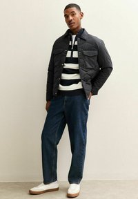 Czarna pikowana kurtka z dwoma kieszeniami na piersi, noszona na czarnym i białym, paskowanym swetrze dzianinowym. Ciemne dżinsy i białe trampki dopełniają stylizację.