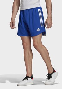Královsky modré sportovní šortky s postranními rozparky, bílými třemi pruhy a logem Adidas na lemu. Nosené s bílými a černými teniskami.