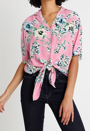 Femme portant un chemisier fleuri rose à manches courtes noué à la taille avec un jean taille haute foncé sur un fond uni.