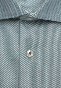Gros plan sur le col d'une chemise bleue à motifs et un bouton argenté cousu avec un fil foncé sur la patte de boutonnage avant.