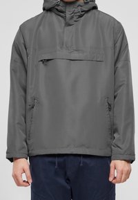 Homme portant un coupe-vent à capuche gris foncé avec poches zippées à l'avant et pantalon bleu marine.