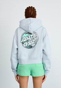 Sweatshirt cropped de cor azul clara, apresentando uma estampa gráfica de "Santa Cruz" com ondas nas costas, combinado com calções verdes.
