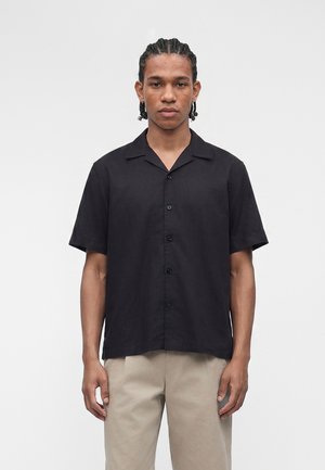 Ung mand med flettet hår iført en sort kortærmet skjorte med knapper og beige bukser, stående mod en ensfarvet hvid baggrund.