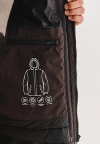Wnętrze brązowej kurtki z kieszenią zapinaną na zamek, na której widnieje biały graficzny wzór kurtki oraz ikony oznaczające odporność na wiatr, tkaninę techniczną i design typu dwa w jednym.