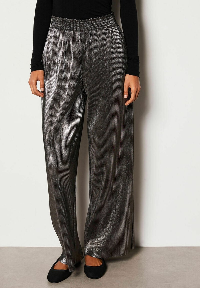 Femme portant un haut noir à manches longues taille haute et un pantalon large argenté plissé brillant avec des fentes latérales ainsi que des chaussures plates noires.