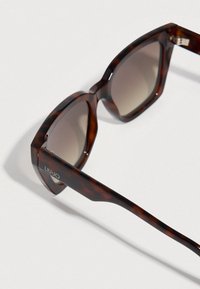 Γυαλιά ηλίου με σκελετό σε σχέδιο χελώνας και τετράγωνο σχήμα, φακούς σε καφέ διαβάθμιση και ασημί διακοσμητικό στο κλαδί με την επωνυμία Liu Jo.