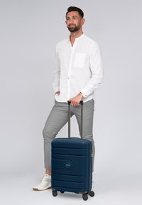 Mann in weißem Hemd und grauen Hosen, der mit einer Hand in der Tasche und mit der anderen den Griff eines blauen Rollkoffers hält.