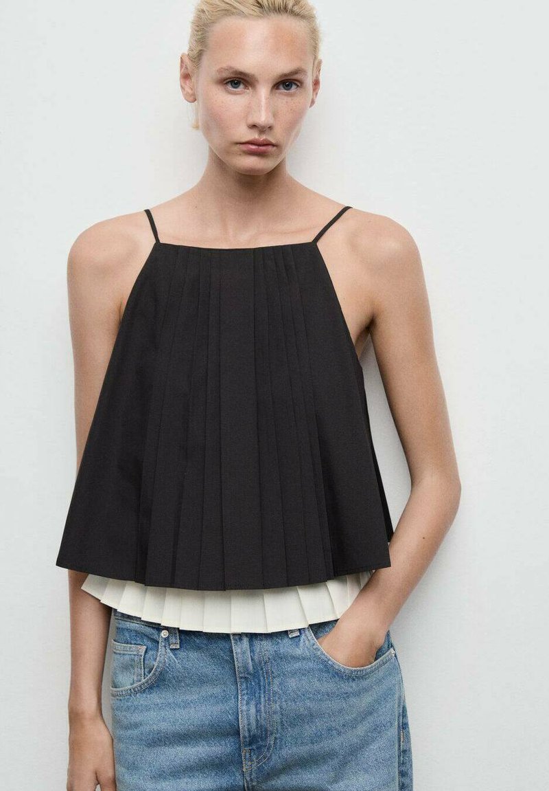 Donna con top nero senza maniche a pieghe sovrapposto a una maglietta bianca e jeans in denim blu, in piedi contro una parete chiara e uniforme.