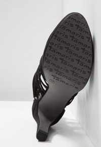 Sandale noire à talon haut inclinée montrant la semelle avec un motif répétitif du logo "tamaris" et une marque de taille 37.
