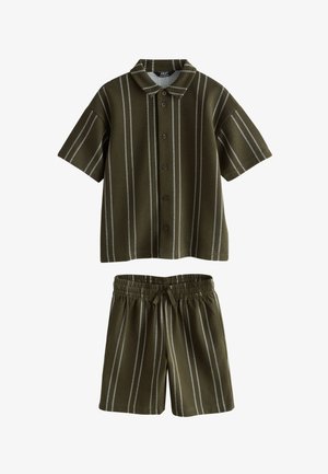 Garçon debout, les mains dans les poches, portant une chemise à manches courtes à rayures verticales vert foncé avec boutons et un short assorti avec des baskets blanches.