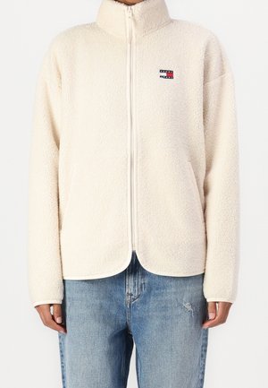 Chaqueta de forro polar color crema con cremallera y cuello alto, con el logo de Tommy Jeans en el pecho, combinada con jeans azul claro desgastados.