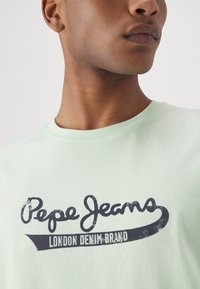 Gaišzaļš kokvilnas T-krekls ar tumši zilā krāsā "Pepe Jeans" logotipu un tekstu "London Denim Brand", klasiskā apaļā kakla izgriezumā.