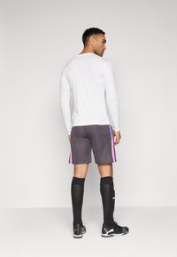 Camiseta atlética blanca de manga larga, pantalones cortos grises con rayas laterales en rosa y púrpura, calcetas negras hasta la rodilla y zapatillas deportivas negras.