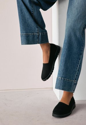Persoon in een paar cropped blauwe jeans en zwarte instapschoenen, met één voet tegen een witte muur steunt op een lichte vloer.