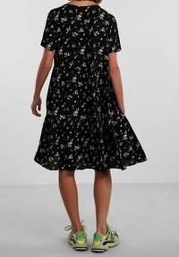 Černé šaty s krátkými rukávy a vrstvovým designem, zdobené květinovým vzorem v světlých barvách. Nošeny s barevnými teniskami.