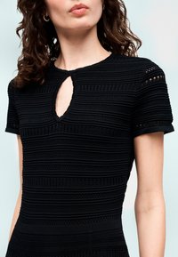 Γυναίκα που φοράει μαύρο κοντομάνικο τοπ με υφή, με μικρό άνοιγμα στο λαιμό και χρυσούς κρίκους σκουλαρίκια, μπροστά σε φωτεινό φόντο.