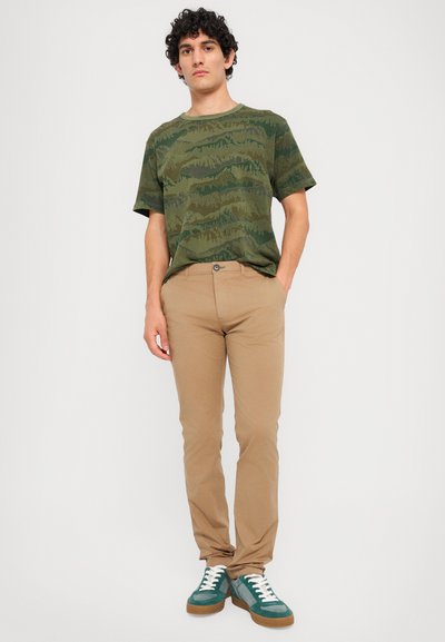 T-shirt verde mimetico con maniche corte, pantaloni aderenti beige e sneakers turchesi con lacci bianchi e suola marrone. Presentazione di un outfit casual.