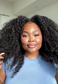 Modell med voluminöst, lockigt svart hår, iförd en ljusblå åtsittande skjorta. Mjuk hy, neutral läppfärg och subtil ögonmakeup.