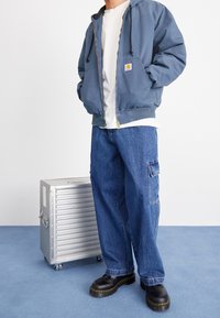 Blauwe bomberjack met capuchon en trekkoord, grijs T-shirt, losse blauwe cargobroek en zwarte platform schoenen. Zilveren koffer in de buurt.