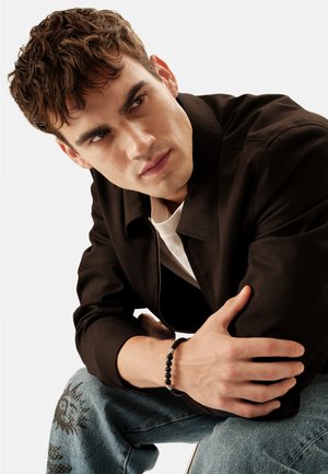 Junger Mann mit lockigem braunem Haar, der eine schwarze Jacke, ein weißes Hemd, ein Perlenarmband und blaue Jeans mit schwarzen grafischen Aufdrucken trägt und sich nach vorne lehnt.