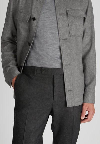 Graue Wollknopfjacke mit Brusttasche, über einem grauen Strickshirt getragen, kombiniert mit dunkelgrauen strukturierten Hosen.