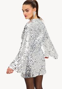 Vestido mini de lentejuelas plateadas con mangas largas de campana y un corte holgado. Presenta un escote redondo y una textura reflectante y brillante.