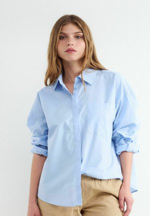 Młoda kobieta o długich jasnobrązowych włosach, ubrana w luźną jasnoniebieską koszulę na guziki z podwiniętymi rękawami oraz beżowe spodnie z fakturą.