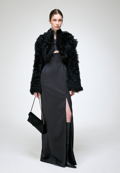 Femme portant une longue robe noire fendue à la cuisse, une veste en fourrure noire texturée, des bottes noires, et tenant un sac à main noir avec une chaîne dorée.