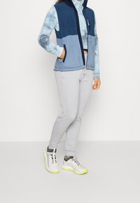 Osoba oblečená v modré fleecové vestě přes dlouhý rukáv s barevným potiskem, světle šedých teplákách a bílých teniskách s neonově žlutými podrážkami.