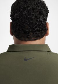 Alyvuogių žalia polo marškinėliai su tekstūruota medžiaga ir juodu Nike logotipu ant nugaros apykaklės. Yra sulankstytos apykaklės dizainas.