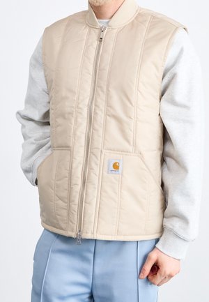 Homme portant une veste matelassée beige zippée par-dessus un sweat-shirt gris clair et un pantalon bleu clair, debout les mains dans les poches.